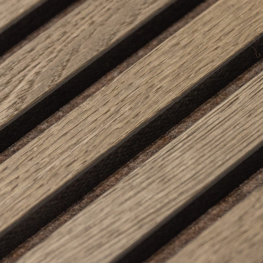 Imagen en primer plano de un panel acústico WoodUpp: listones paralelos de madera grisácea con veta visible y acabado mate, separados por ranuras negras rellenas de fieltro acústico marrón oscuro. Producto WoodUpp diseñado para revestimiento decorativo y absorción sonora en paredes o techos.