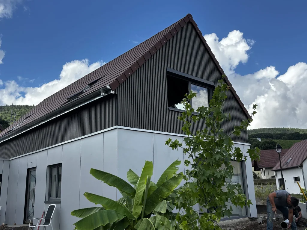 Fachada de vivienda con marquesina triangular revestida con listones verticales WoodUpp de tono oscuro en el piso superior y paneles lisos WoodUpp color gris claro en la planta baja; ventana rectangular, canalón metálico, planta tropical en primer plano y operario trabajando junto al acceso.