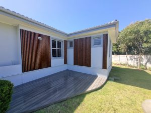 Fachada de casa con jardincillo y elementos WoodUpp: paneles verticales de listones de madera teñida instalados como celosías decorativas y una tarima WoodUpp gris de lamas compuestas, junto a paredes blancas, ventanas y césped, mostrando contraste entre madera cálida y diseño exterior moderno.