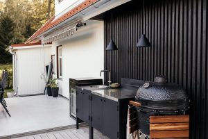 Espacio exterior WoodUpp: cocina modular negra con encimera de acero, fregadero redondo y armarios con puertas, mini nevera integrada y parrilla kamado con estantes laterales de madera. Fondo de listones negros, lámparas colgantes cónicas y terraza amplia junto a la casa blanca.