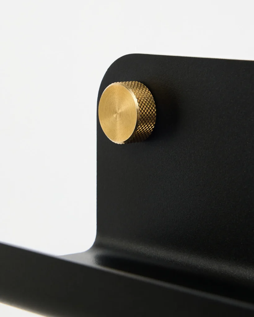 Detalle de producto WoodUpp: placa metálica negra con acabado mate y borde redondeado y un pequeño pomo/tornillo embellecedor de latón dorado con textura cuadriculada (knurled) y acabado de precisión. Diseño elegante, minimalista y robusto, parte de un soporte o estante de pared WoodUpp.