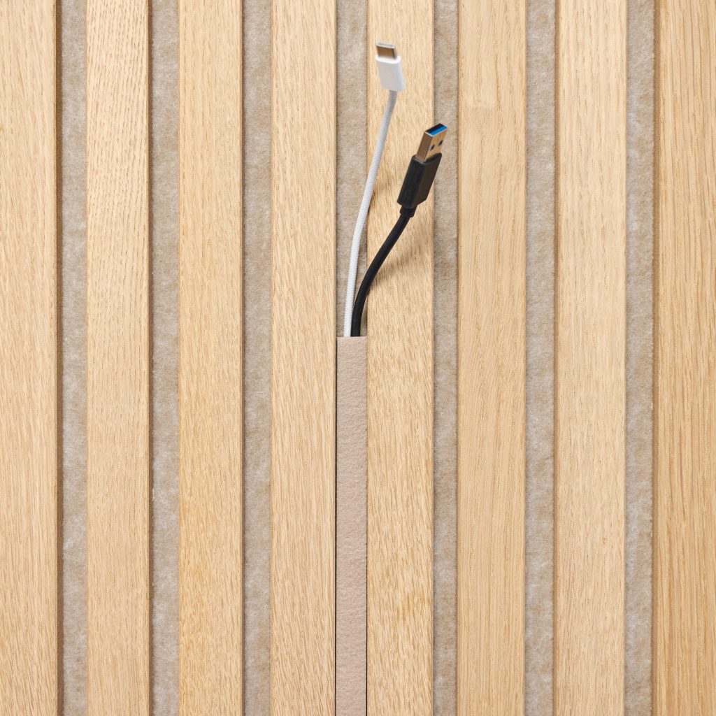 Panel WoodUpp de listones de madera clara con fieltro gris entre las lamas, mostrando una ranura vertical para gestión de cables. Se ven dos cables saliendo: uno blanco con conector Lightning y otro negro con USB-A azul, resaltando la funcionalidad y acabado natural del producto.