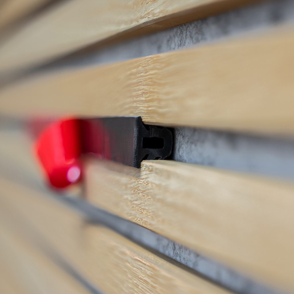 Primer plano de lamas horizontales de madera clara WoodUpp, con el perfil de su sistema de fijación WoodUpp: un clip negro encajado entre las lamas y un elemento de bloqueo plástico rojo visible al fondo, mostrando textura de la madera y el montaje del panel.