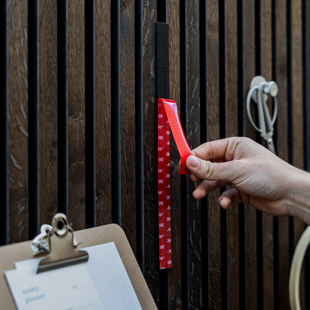 Panel de lamas de madera WoodUpp en tonos oscuros con un perfil vertical magnético WoodUpp fijado; una mano despega la tira adhesiva roja 3M del accesorio. Se aprecian además un portapapeles y una cuerda con gancho colgados en las lamas.