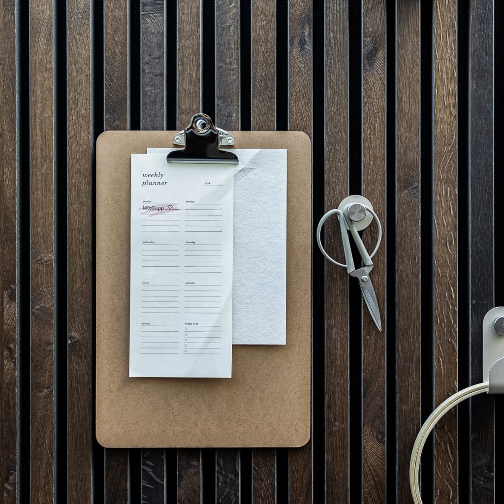 Panel de lamas verticales WoodUpp en tonos madera oscura, con un portapapeles de madera MDF sujetando un weekly planner que tiene "WoodUpp" escrito, y accesorios magnéticos WoodUpp: un gancho circular con tijeras colgadas y un soporte para cable visible a la derecha.