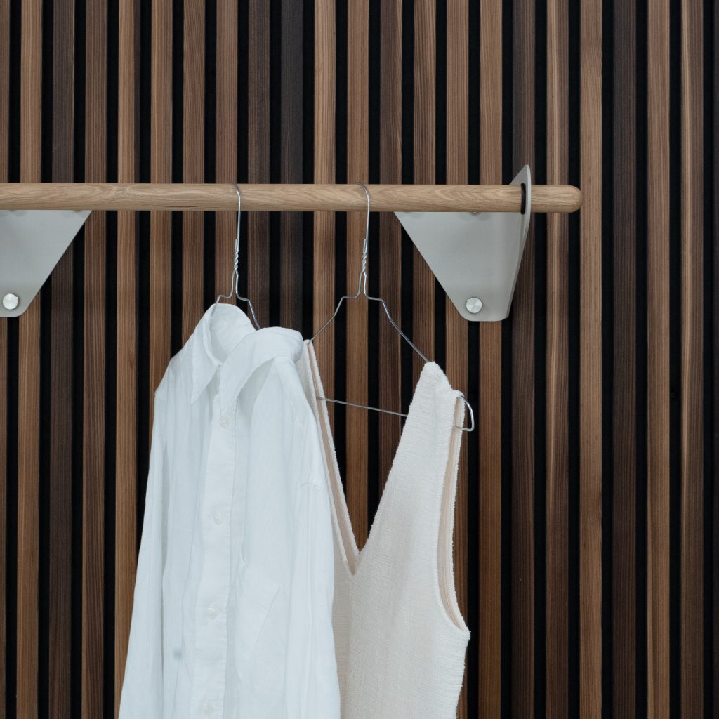 Panel de listones verticales WoodUpp en tonos nogal y roble, con una barra colgadora de madera WoodUpp sujeta por soportes triangulares metálicos blancos. Sobre la barra cuelgan perchas de alambre con una camisa blanca y un top beige, en primer plano.