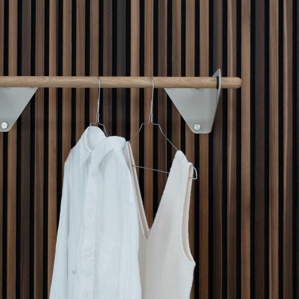 Panel de listones verticales WoodUpp en tonos nogal y roble, con una barra colgadora de madera WoodUpp sujeta por soportes triangulares metálicos blancos. Sobre la barra cuelgan perchas de alambre con una camisa blanca y un top beige, en primer plano.