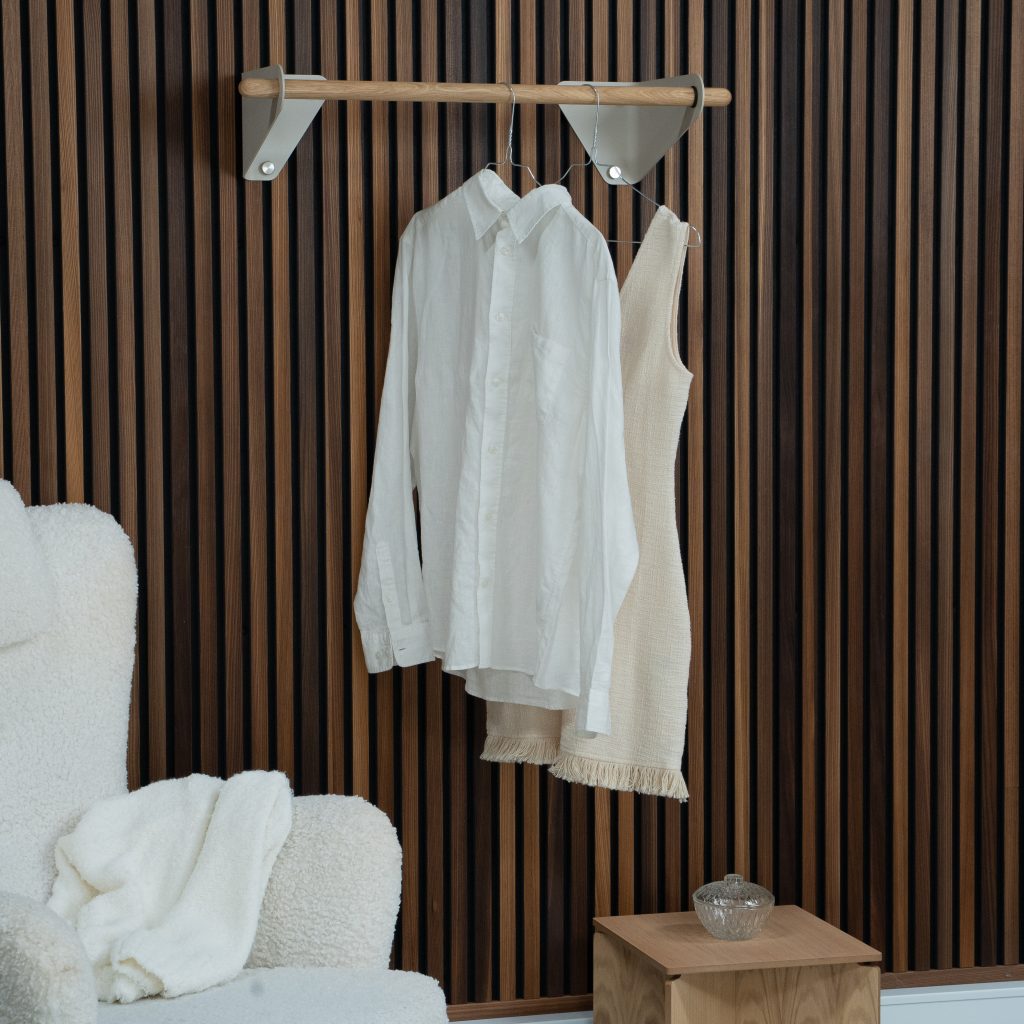 Barra de madera WoodUpp con soportes triangulares de pared WoodUpp montada sobre listones verticales de madera; colgada una camisa blanca y un vestido beige en perchas metálicas. A la izquierda sillón blanco de borreguito con manta y a la derecha mesita de madera con recipiente de cristal.