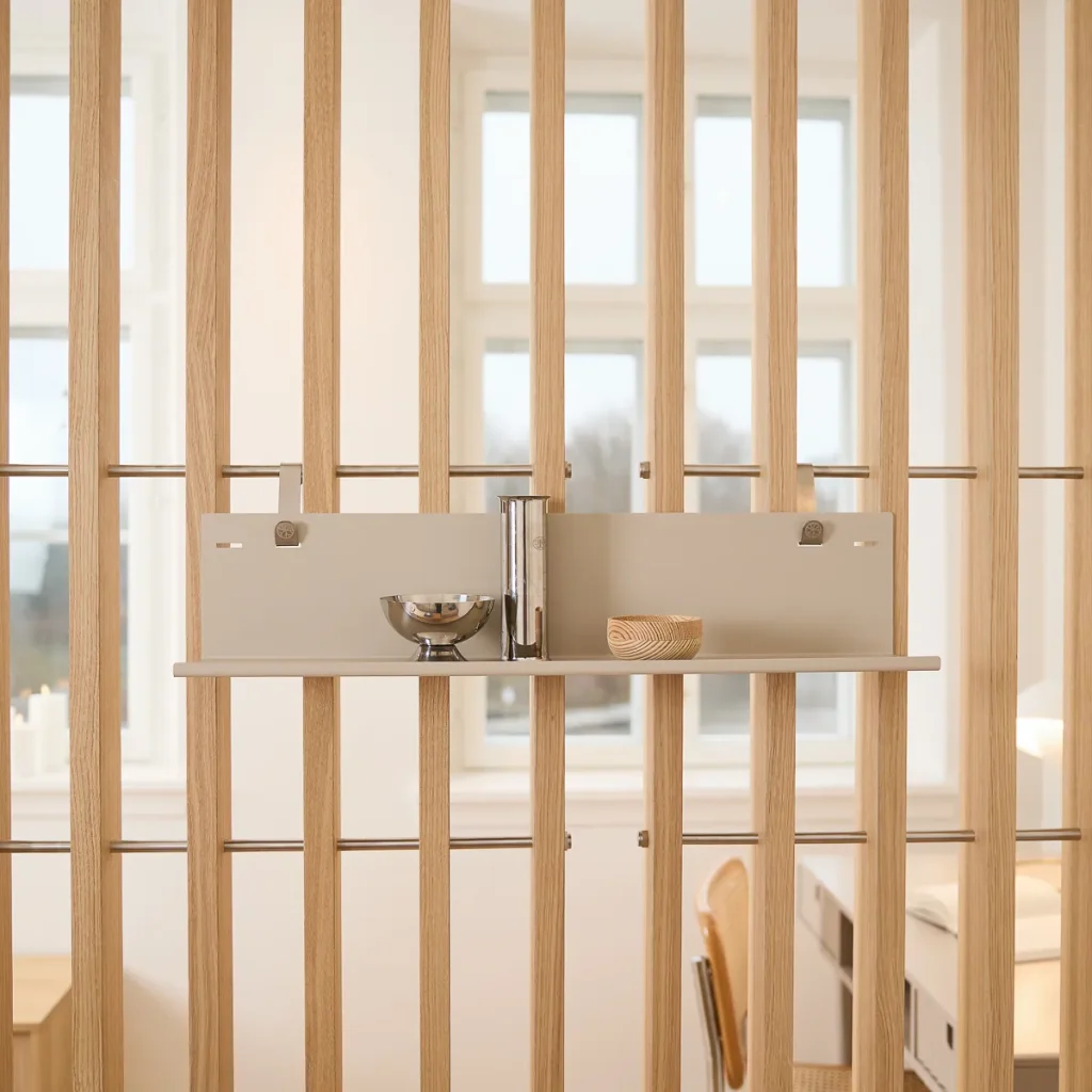 Separador de ambiente de lamas verticales de madera clara WoodUpp con barras metálicas horizontales, acompañado de un panel modular beige y estante colgante WoodUpp fijado con clips metálicos. En el estante: un vaso cilíndrico cromado, un cuenco metálico y un pequeño cuenco de madera.