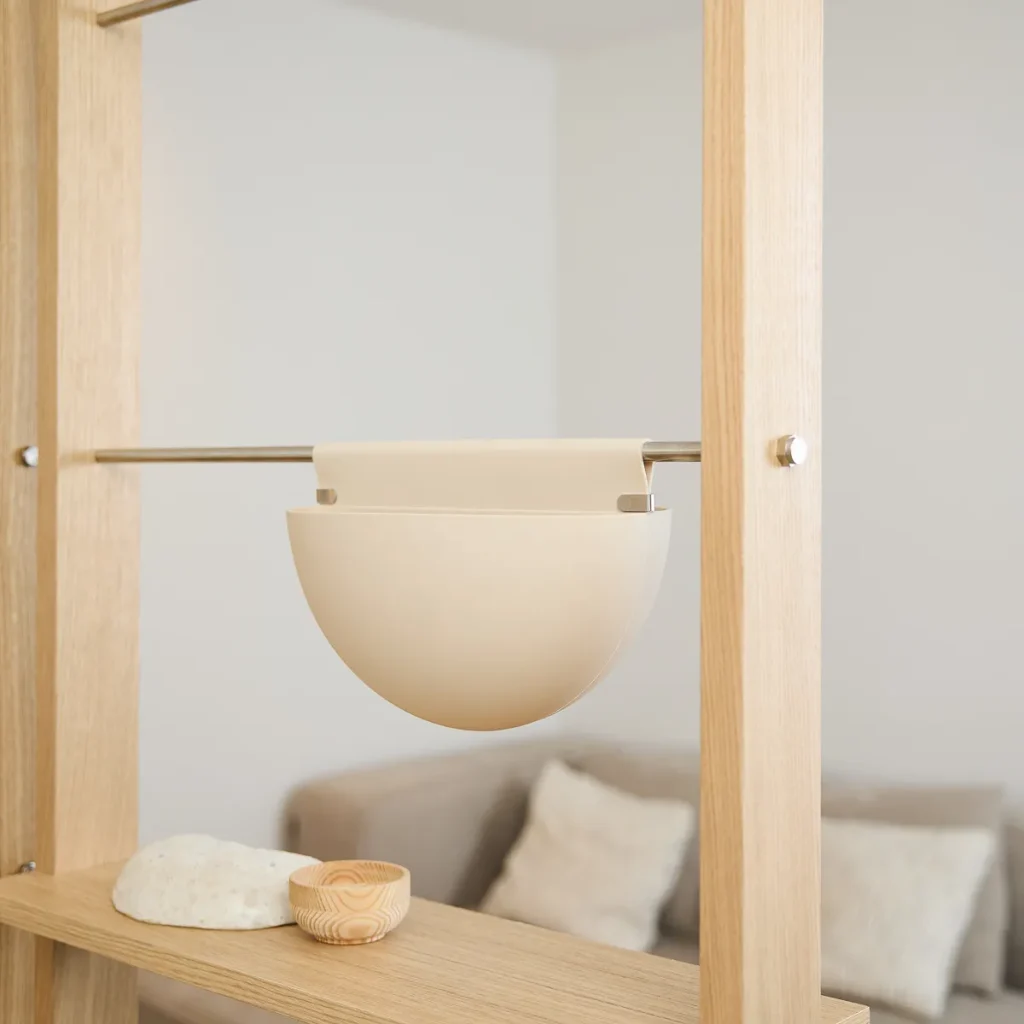Estantería modular WoodUpp en madera clara con barra metálica, mostrando una maceta colgante WoodUpp semiesférica beige fijada al soporte y un estante bajo con pequeño cuenco de madera y piedra decorativa; fondo neutro con sofá y cojines desenfocados, iluminación cálida y minimalista.