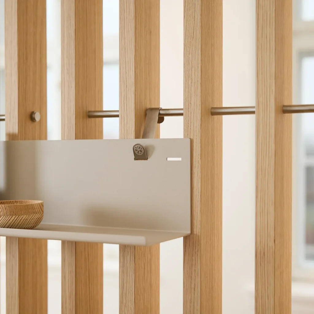 Panel separador vertical de lamas de roble WoodUpp con barra metálica; estante colgante metálico beige de WoodUpp sujeto con gancho y un pequeño cuenco de madera sobre la repisa. Acabado natural y diseño modular, mostrando herrajes y unión discreta, elegante y funcional.