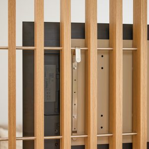 Panel de lamas verticales de madera clara WoodUpp con barras horizontales metálicas; detrás se aprecia la parte trasera de un televisor sujeto al soporte metálico regulable de WoodUpp, con anclajes y ranuras visibles. Imagen en primer plano del detalle del montaje y estructura.