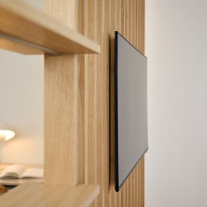 Panel vertical de listones de madera clara WoodUpp con estantes integrados y televisor de pantalla plana montado de forma discreta; el sistema modular muestra vetas longitudinales y acabado natural, creando un mueble divisor funcional que soporta estanterías y la pantalla empotrada, ideal para espacios minimalistas.