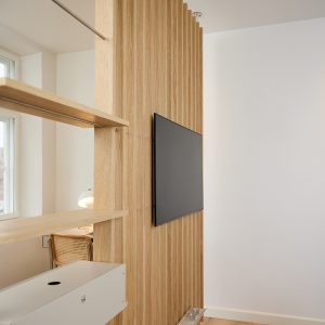 Panel divisor vertical de lamas de roble WoodUpp con televisor montado, estantes flotantes WoodUpp integrados y riel metálico; se aprecia una silla con respaldo de rejilla, una lámpara de sobremesa y una ventana en un espacio luminoso de diseño minimalista.