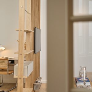 División de ambiente WoodUpp: estantería modular de madera de roble con lamas verticales, baldas abiertas y caja organizadora blanca integrada; soporte de TV montado en la estructura y pequeña bandeja inferior para mandos. Ambiente luminoso con escritorio, silla de rejilla y decoración minimalista.