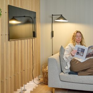 Interior acogedor con sofá y lámpara, mostrando un revestimiento de pared WoodUpp: paneles de lamas de madera claras instaladas verticalmente como listones decorativos, con soportes metálicos en la base; televisión montada en el panel y una mujer leyendo en el sofá.