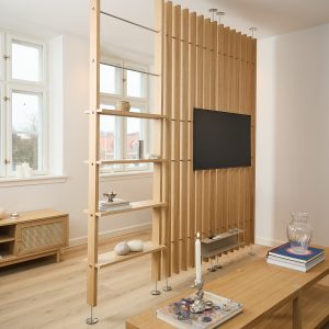 Divisor WoodUpp de listones verticales en roble claro con soporte para televisor y estanterías integradas; panel separa espacios, fijaciones metálicas visibles y baldas abiertas con objetos decorativos (libros, vela, cuencos). Diseño funcional que actúa como mueble multimedia y estantería en salón luminoso.