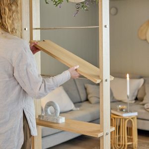 Persona colocando un estante de la estantería modular WoodUpp: postes verticales de roble claro con tornillería metálica y barras horizontales cromadas, y estantes desmontables de madera maciza tipo tablón. Diseño abierto y regulable, ideal como separador de ambientes en salón.