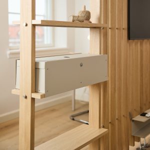 Divisor estantería de madera clara WoodUpp con herrajes visibles; estante sostiene una caja metálica beige de WoodUpp para organización de cables con ranura superior y orificio de ventilación. Encima, un ovillo de cuerda decorativo. Diseño modular y acabado natural, estilo minimalista y funcional.