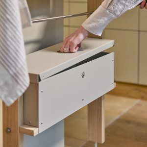 Mano abriendo la tapa de un cajón metálico blanco de WoodUpp, integrado en una estructura modular de madera clara con barra metálica para colgar ropa. El producto muestra tornillos visibles, un orificio redondo en la tapa y un diseño minimalista y funcional.