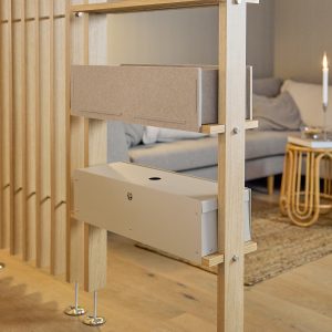 Panel divisor modular WoodUpp de listones de roble con bastidor vertical, que sostiene dos productos WoodUpp: una caja organizadora de fieltro marrón colocada en una repisa y una caja metálica beige para gestión de cables y electrónica. Ambiente de salón con sofá y vela al fondo.