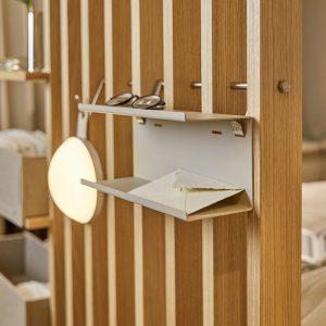 Panel separador de lamas de madera WoodUpp con estantería metálica modular WoodUpp colgada: bandeja inclinada para sobres y repisa superior para gafas. A la izquierda se aprecia una lámpara redonda suspendida y el ambiente del mobiliario, todo en tonos cálidos y diseño minimalista.