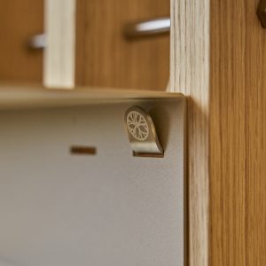 Detalle de WoodUpp: panel vertical de madera clara tipo roble y placa metálica mate con ranuras, junto a un pequeño gancho metálico WoodUpp con símbolo circular grabado. Se aprecian vetas de la madera y el acabado texturizado del metal en primer plano.