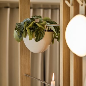 Panel separador de madera WoodUpp con riel metálico que sostiene una maceta colgante blanca WoodUpp en forma de media esfera, llena de una planta de hojas verdes colgantes. En primer plano, vela encendida y luz redonda difusa complementan la escena.