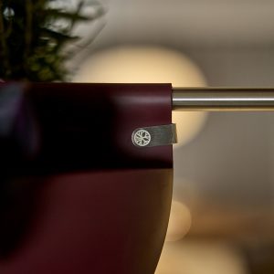 Macetero colgante burdeos de WoodUpp sujeto a una barra metálica dorada mediante un clip metálico con el logo circular de WoodUpp; se aprecia la parte superior de una planta verde desenfocada y el diseño minimalista y mate del macetero en primer plano.