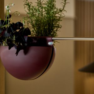 Jardinera colgante WoodUpp granate en forma semicircular sujeta a una barra metálica con clip WoodUpp visible; contiene plantas verdes y hojas púrpuras (Oxalis) y hierbas tipo romero. Diseño moderno de maceta metálica suspendida, acabado mate y sujeción minimalista de acero.