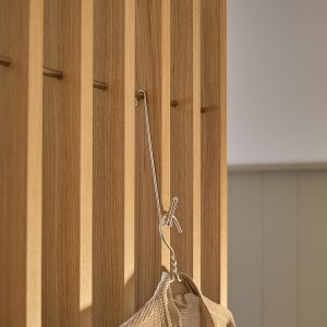 Panel de listones verticales de roble de WoodUpp con acabado natural, mostrando clavijas/percheros cilíndricos de latón WoodUpp insertados entre las ranuras. Un gancho metálico sostiene una bolsa de tejido beige, destacando la modularidad y la estética minimalista del sistema WoodUpp.