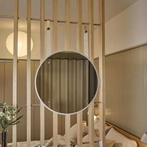 Panel separador de listones de roble claro de WoodUpp instalado con varillas metálicas, con un espejo redondo colgante WoodUpp de marco blanco suspendido mediante correas ajustables beige y hebillas metálicas; fondo con cama, cortina y lámpara esférica, ambiente cálido y minimalista.