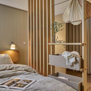 División de ambiente WoodUpp: estantería modular de madera clara con barras metálicas, baldas y dos cajas organizadoras de fieltro gris con ropa, colocada junto a cama. En la escena se ven lámpara de pared, cabecero de madera, revista abierta y planta decorativa sobre la balda.