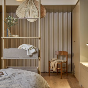 Un dormitorio moderno presenta el separador de ambientes de roble de WoodUpp. Esta estructura de madera incluye estantes con decoración y prácticas cajas de fieltro beige. Divide el espacio entre la cama con colcha gris y un área con una silla de madera y cortinas.