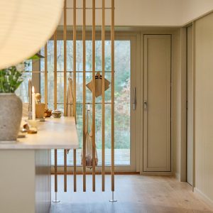 Biombo de listones verticales de roble claro de WoodUpp, instalado como separador entre cocina y ventanal; incluye barra metálica y ganchos para colgar paños, bolsita de malla y tablas de madera. Ambiente luminoso y minimalista con encimera clara y detalles decorativos sobre la isla.