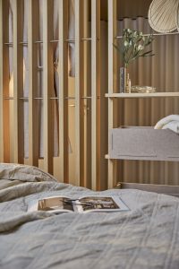 Detalle de dormitorio con los paneles de listones de roble de WoodUpp, barra metálica para colgar ropa y estantería modular WoodUpp con cajas de fieltro beige. Sobre la cama, ropa de cama neutra y un libro abierto completan la escena acogedora.