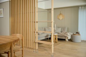 Panel divisor de ambiente WoodUpp formado por lamas verticales de roble claro fijadas con tirantes metálicos, acompañado de una estructura escalonada-estantería WoodUpp integrada con baldas de madera y patas metálicas. Sala minimalista con sofá gris, alfombra de fibras naturales y vela encendida.