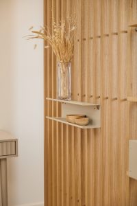 Paneles de lamas verticales de madera WoodUpp en tono roble, con textura acanalada, y dos estantes metálicos WoodUpp color beige montados a la pared. En ellos hay un jarrón de cristal con flores secas y dos pequeños cuencos de madera, creando un rincón decorativo minimalista.