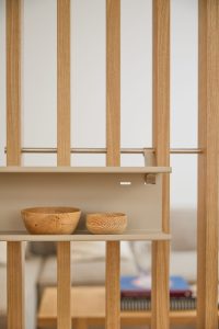 Separador de ambientes WoodUpp con listones verticales de madera clara y riel metálico, acompañado del estante modular beige WoodUpp enganchado al panel; en la balda, dos cuencos de madera natural con vetas visibles, acabado liso, aspecto artesano y diseño minimalista.