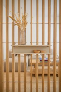 Separador de ambiente de lamas verticales de roble WoodUpp, con barras metálicas horizontales; estantería flotante modular beige de WoodUpp anclada al separador, de dos niveles y líneas limpias. Sobre ella, un jarrón alto de cristal con ramas secas y dos cuencos de madera decorativos.