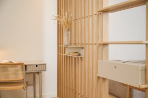 Panel separador vertical de lamas de roble WoodUpp con una estantería flotante beige y un cajón modular WoodUpp montados, mostrando un estante metálico pequeño con cuencos de madera; ambiente nórdico con escritorio y silla de rejilla en tonos cálidos y paneles abiertos que separan espacios interiores.