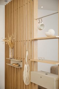 Panel separador de lamas de roble WoodUpp con estanterías integradas: baldas de madera flotantes, estantes metálicos beige, cajón metálico y ganchos modulables. En la imagen aparecen una taza colgada, una bolsa de malla, un jarrón con flores secas y pequeños recipientes decorativos.