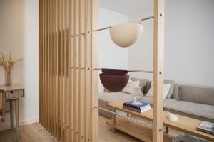 Panel separador WoodUpp de listones verticales de madera clara con varillas metálicas, mostrando dos maceteros colgantes WoodUpp semiesféricos —uno beige y otro burdeos— sujetos a las barras. Escena en salón minimalista con sofá, mesa de madera y detalles decorativos luminoso.