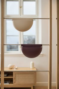 Estantería de madera clara WoodUpp con dos contenedores colgantes semicirculares de la marca: uno beige y otro burdeos oscuro, suspendidos en barras metálicas. Los recipientes, de diseño minimalista y curvado, sirven como organizadores decorativos frente a una ventana y un mueble bajo.