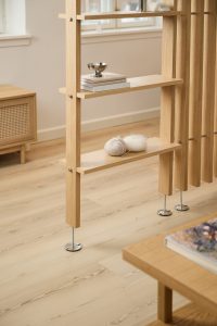 Separador modular de WoodUpp en madera de roble claro con estantes integrados y listones verticales; soportes metálicos regulables. En los estantes se ven libros y un cuenco metálico, y ovillos de lana con agujas. También aparece una mesa baja y un mueble auxiliar a juego.