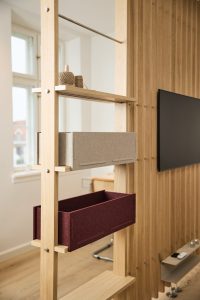 Panel separador WoodUpp de madera clara tipo estantería vertical con estantes estrechos; incluye dos cajas organizadoras de fieltro WoodUpp, una gris claro y otra granate, encajadas en repisas, y un pequeño rollo de cuerda sobre la balda superior, en salón luminoso.