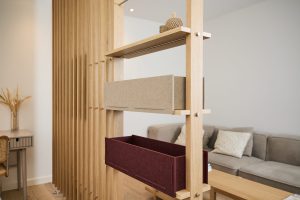 Separador-estantería modular de madera WoodUpp con listones verticales de roble y estantes; incluye dos cajas organizadoras de fieltro WoodUpp (beige y burdeos) y pequeño estante superior con objetos decorativos. Ambientación de salón y escritorio al fondo, diseño minimalista y funcional.