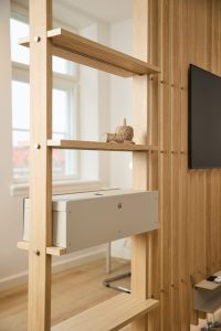 Divisor estantería WoodUpp en roble claro, con largueros verticales y baldas finas, mostrando una caja portacables metálica beige de WoodUpp alojada en una balda y dos rollos de cordel decorativos; al fondo lamas de madera y televisor montado, con detalles metálicos y tornillería vista.