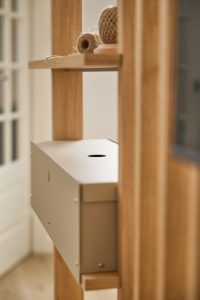 Detalle de una estructura WoodUpp: un armazón vertical de madera clara con una caja metálica beige montada con abertura circular, y un estante superior con rollos de cuerda de yute. Muestra la calidad de la madera y el diseño modular de los productos WoodUpp.