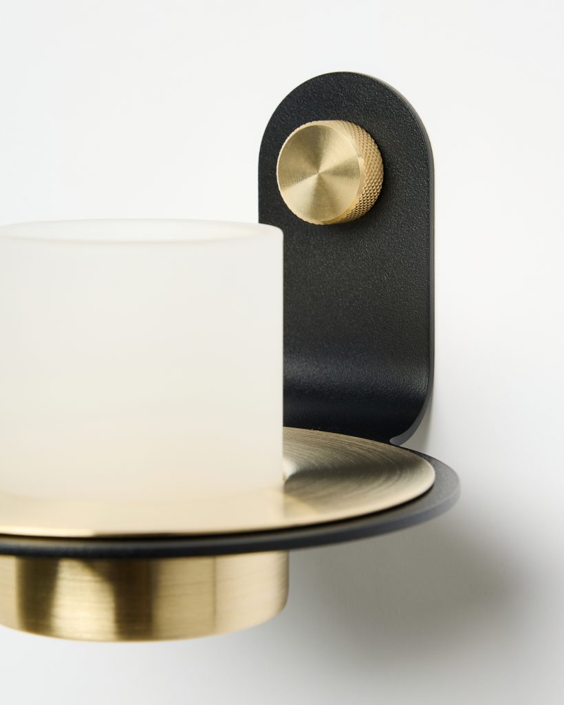 Detalle de un aplique mural WoodUpp: soporte curvado negro mate, plato y casquillo de latón cepillado, perilla estriada de latón dorado y pantalla cilíndrica de vidrio esmerilado blanco. Diseño minimalista y materiales metálicos que combinan negro y latón, mostrado en vista cercana.